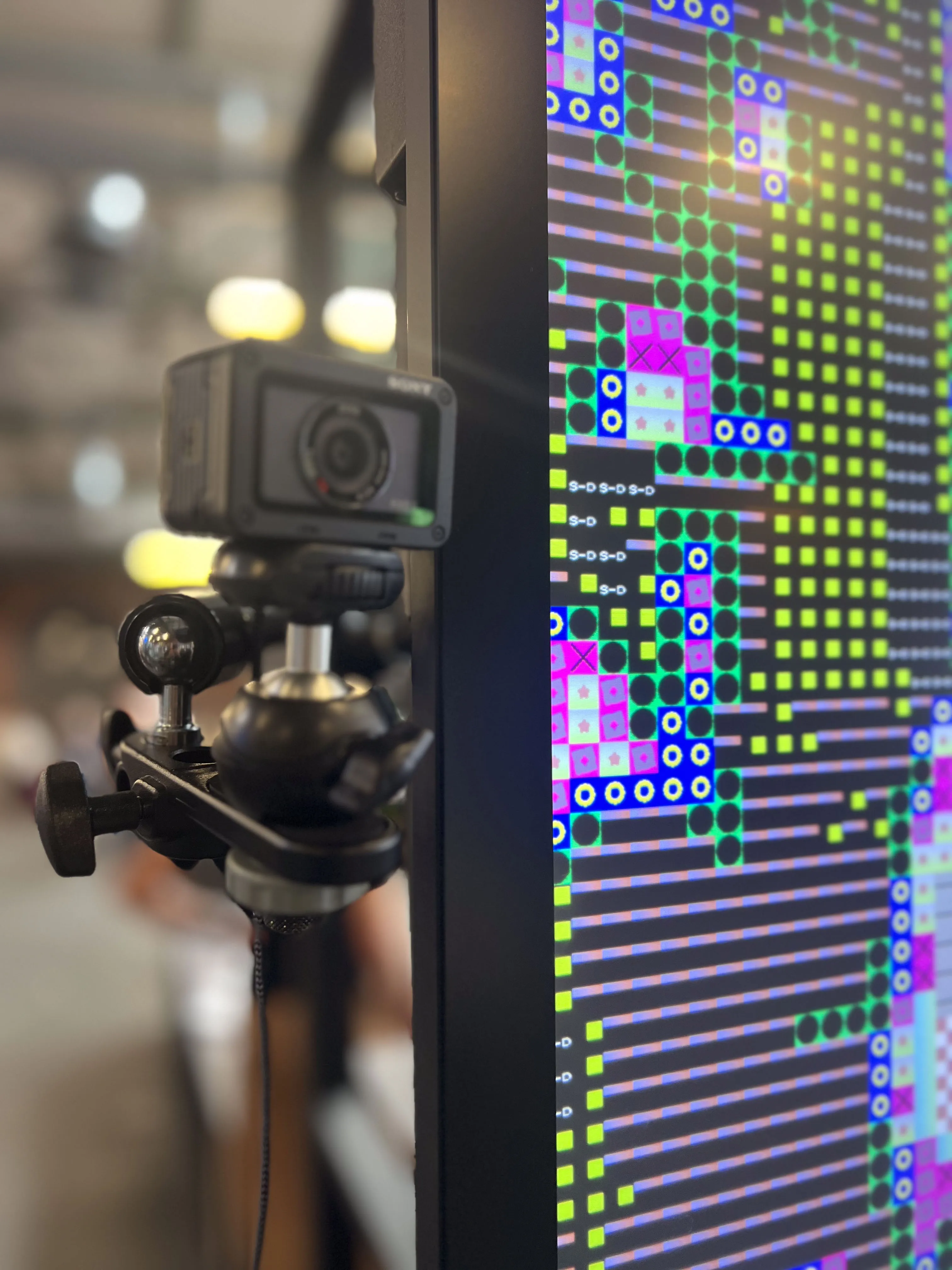 Nahaufnahme des Hardware-Setups — Webcam auf Stativ vor dem Bildschirm mit laufender generativer Pixeldarstellung